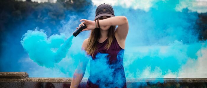 A stylish woman surrounded by bright blue smoke outdoors, creating a dynamic fashion statement.